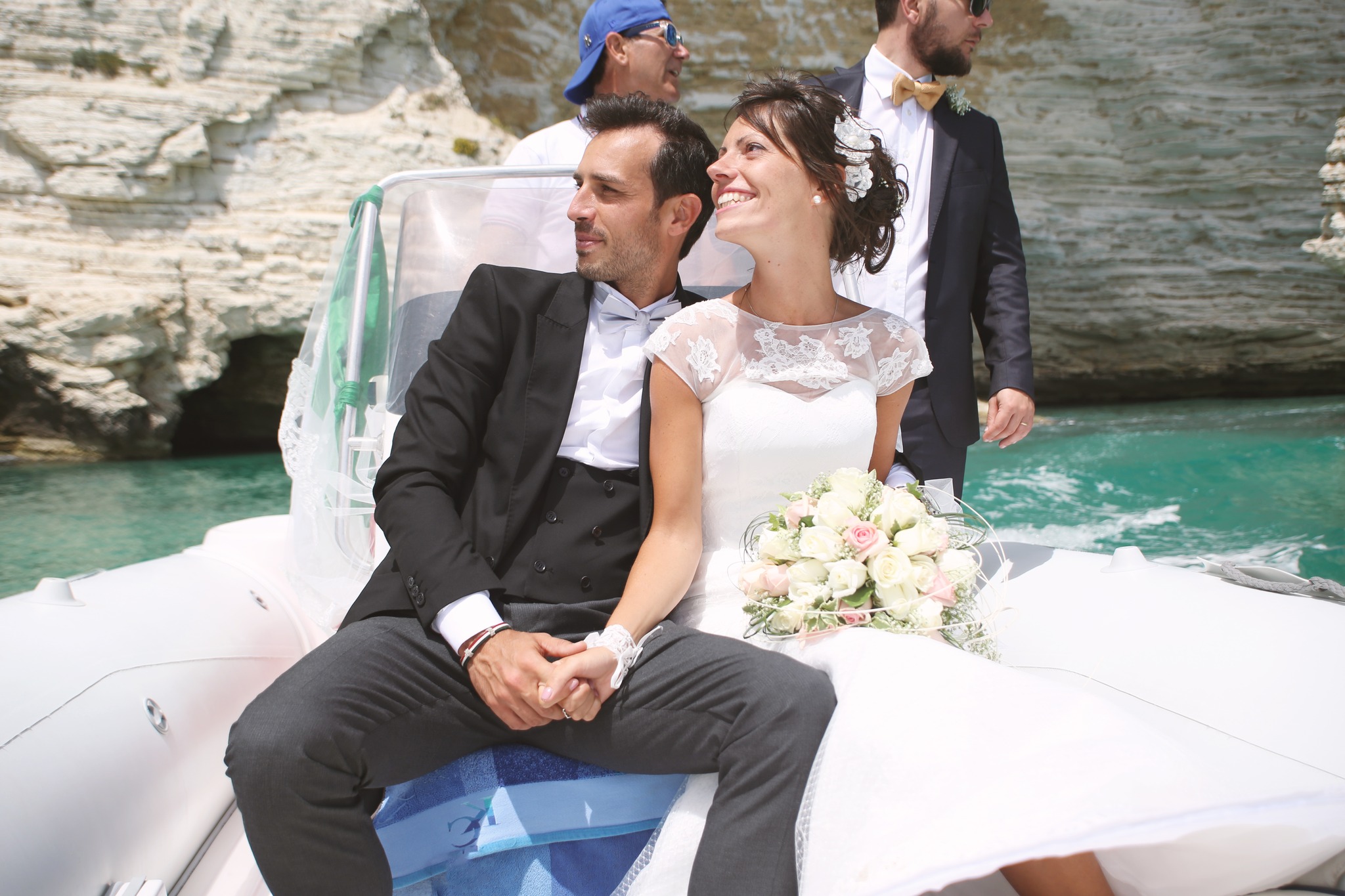 Sposi mano nella mano su un gommone bianco con bouquet di rose, mare turchese e scogliera calcarea, foto RC Studio